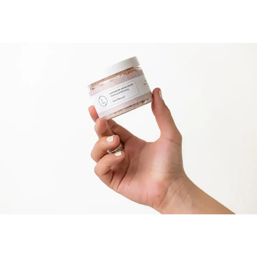 A clear glass jar with a white lid and label, containing lavender bath salts in pinkish-purple granules.