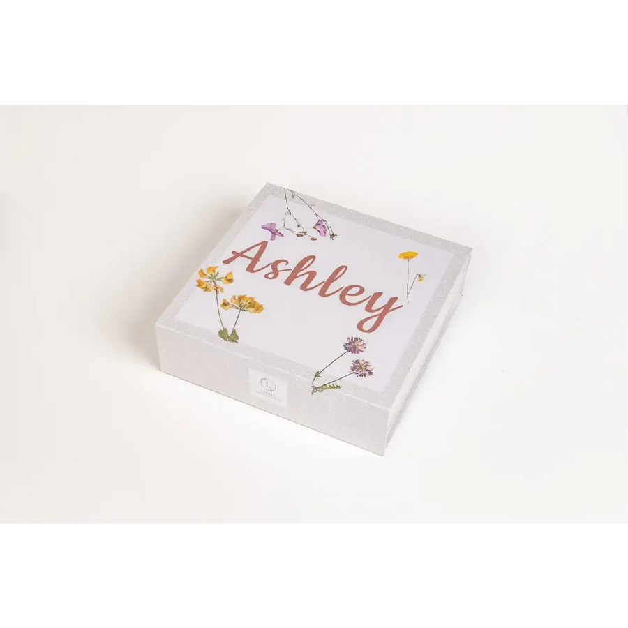 A white square gift box adorned with delicate pressed flowers in yellow, purple, and pink hues around the name ’ashley’ written in a warm terracotta cursive script.