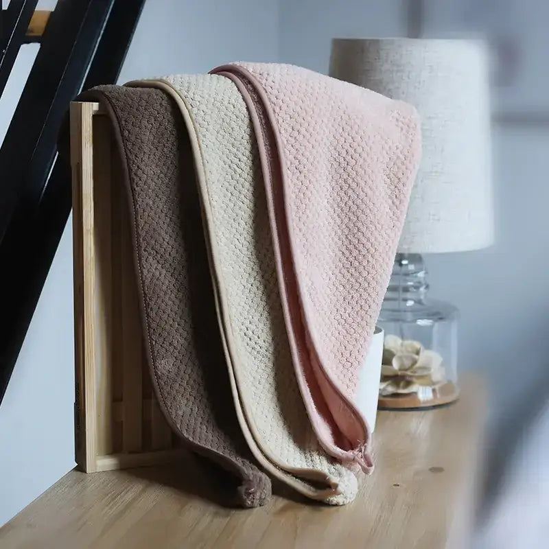Three soft, textured microfiber towels in muted brown, beige, and pink tones are neatly arranged within a light wooden tray.