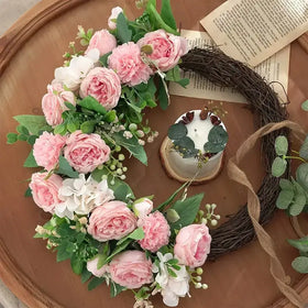 A delicate pink and white floral wreath adorned with lush greenery, crafted from artificial materials to mimic fresh blossoms.