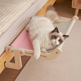 A fluffy cream and brown cat lounges comfortably in a pink hammock-style bed with light wood frame.