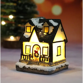 A warmly lit miniature christmas house figurine with a black shingled roof dusted in white snow, glowing yellow interior lights, and a festive wreath adorning the front door.