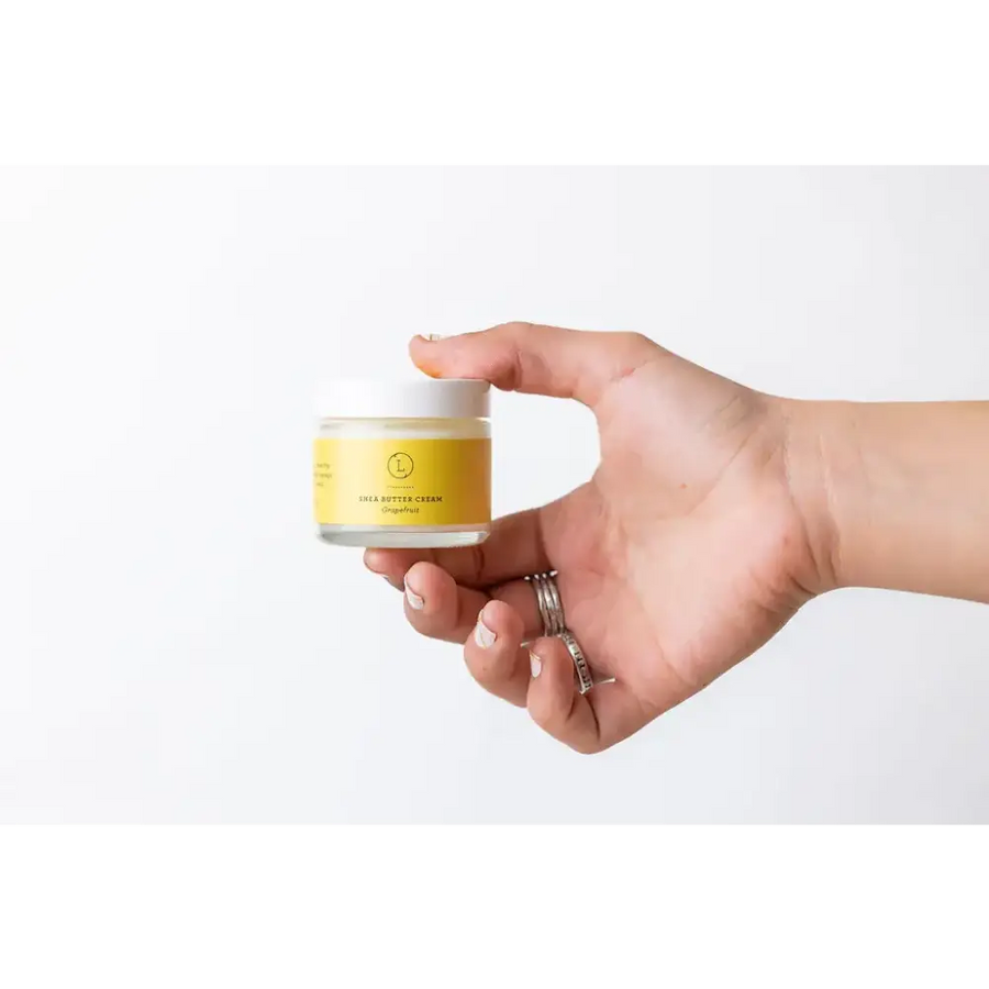 A small, clear glass jar with a white lid and a vibrant yellow label that reads ’shea butter cream’ by the brand lush.