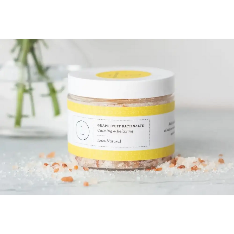 A jar of grapefruit bath salts with a white lid and yellow label, containing pinkish-orange crystalline salt.