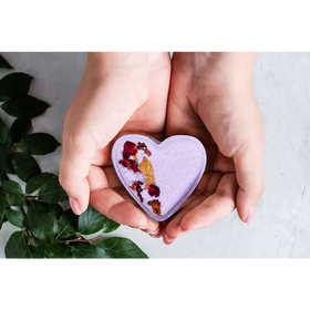 A lavender heart-shaped bath bomb with dried rose petals and gold leaf embedded in its surface.