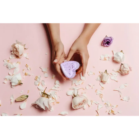 A lavender heart-shaped bath bomb with a speckled texture, held gently in two hands against a soft pink background scattered with rose petals and flowers.