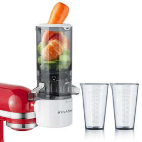 White kitchtree juicer with transparent cylindrical chamber filled with vegetables and fruit, accompanied by two clear measuring cups.