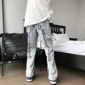 Light-wash denim jeans featuring bold purple script lettering down the outer leg and frayed, multi-colored hem detailing.