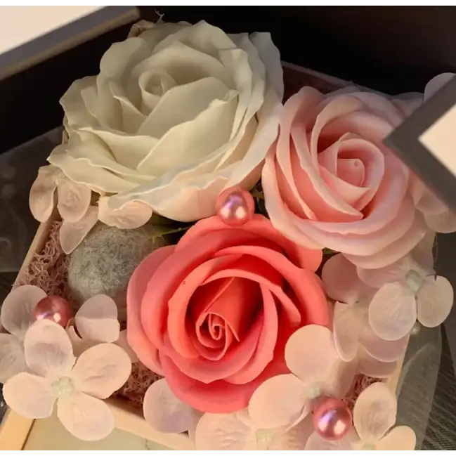 A delicate arrangement of three artificial roses in soft cream, blush pink, and coral hues nestled among tiny white flowers and pearl-like accents.