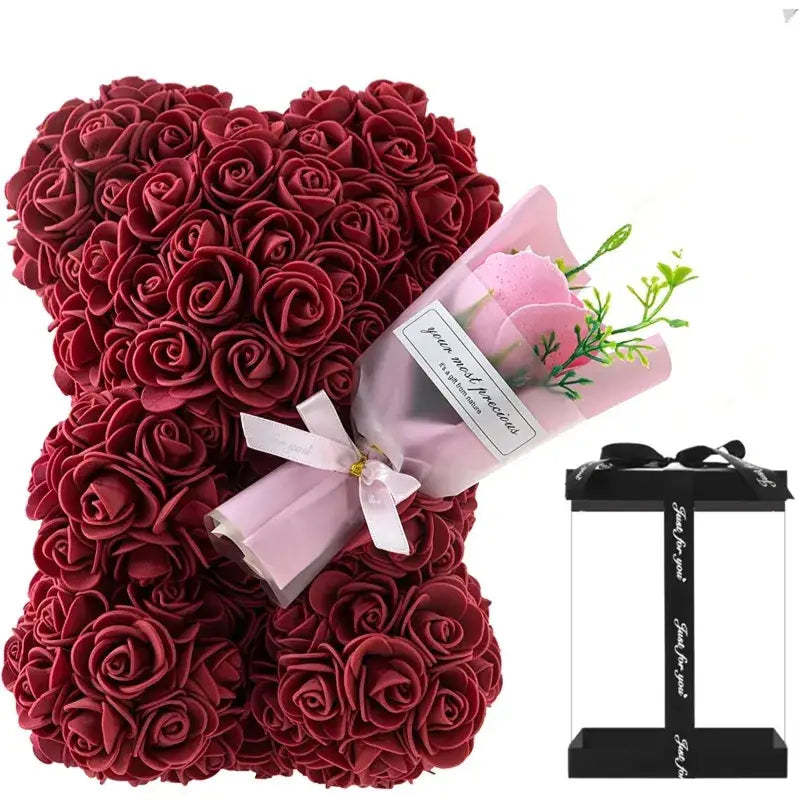 A plush teddy bear crafted from dense clusters of deep red artificial roses, holding a small bouquet wrapped in pink paper with a white ribbon.