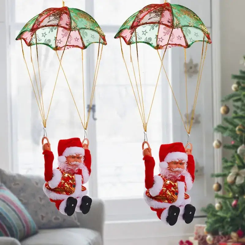 Two festive christmas-themed santa claus figurines, each suspended by a decorative parachute with green and pink patterned fabric, wearing red suits adorned with gold glittery accents.