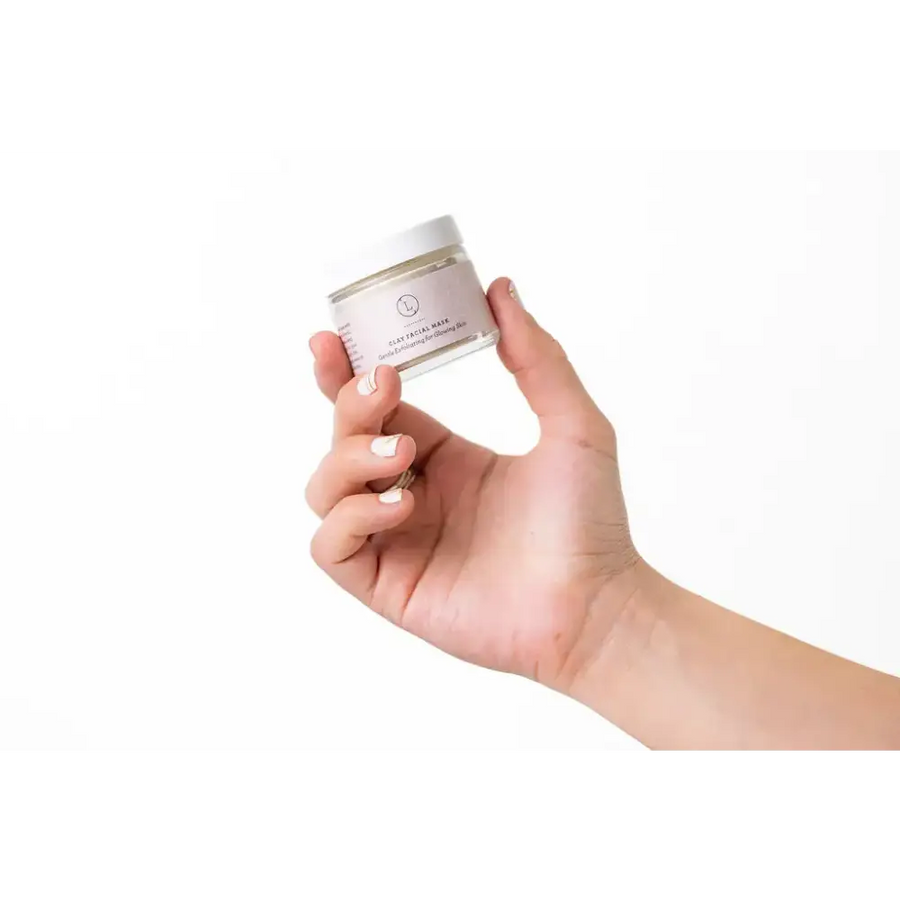 A clear glass jar with a white lid and minimalist label, holding a clay facial mask.