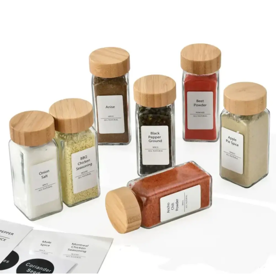 Set of clear glass spice jars with natural wood lids, each labeled with white stickers and containing different colored seasonings.