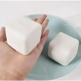 A smooth, off-white, cube-shaped bar of soap with rounded edges rests in a hand beside another similar bar on a light blue plate.