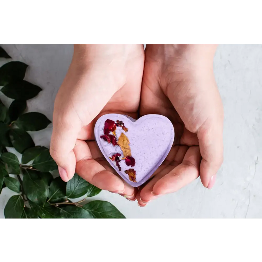 A lavender heart-shaped bath bomb with dried rose petals and gold leaf embedded in its surface.