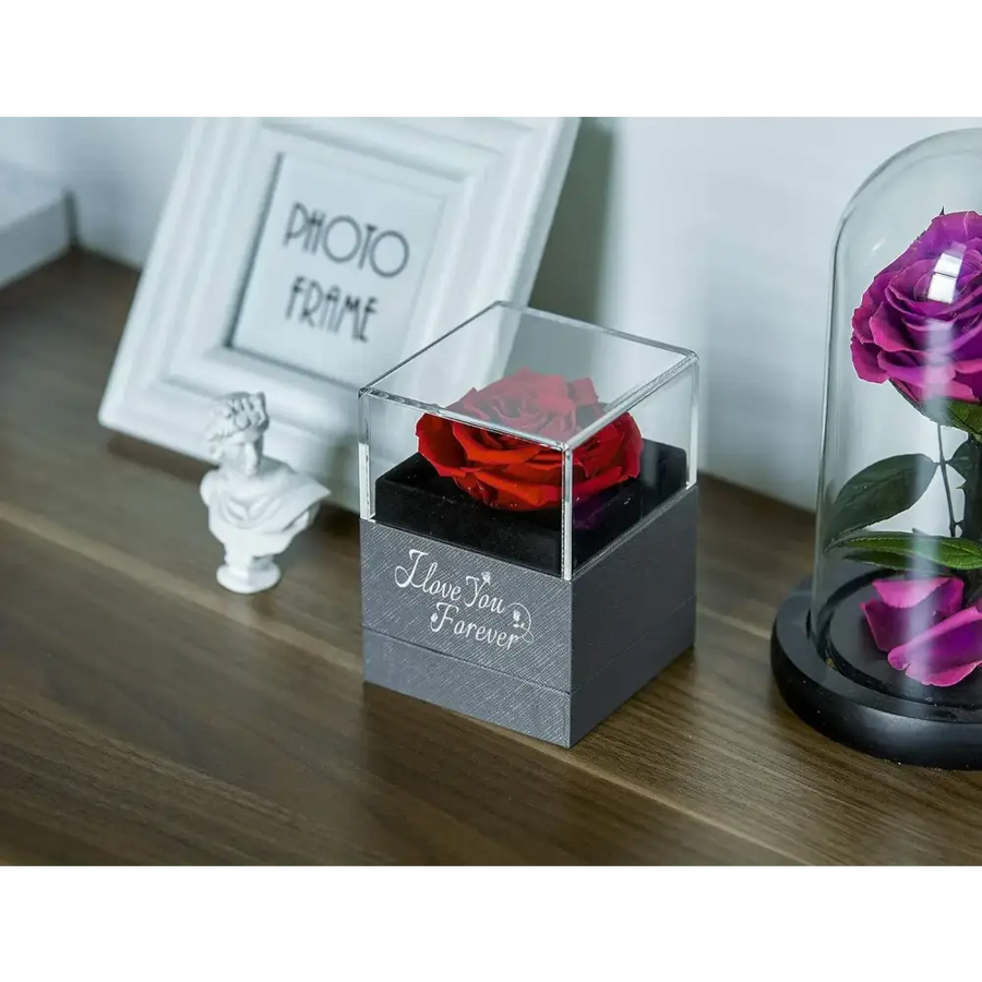 A vibrant red preserved rose nestled inside a clear acrylic cube with a textured gray base engraved with ’i love you forever.’