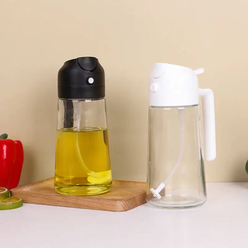 Clear glass oil dispenser with black spray nozzle and yellow liquid, beside an empty one with white handle.