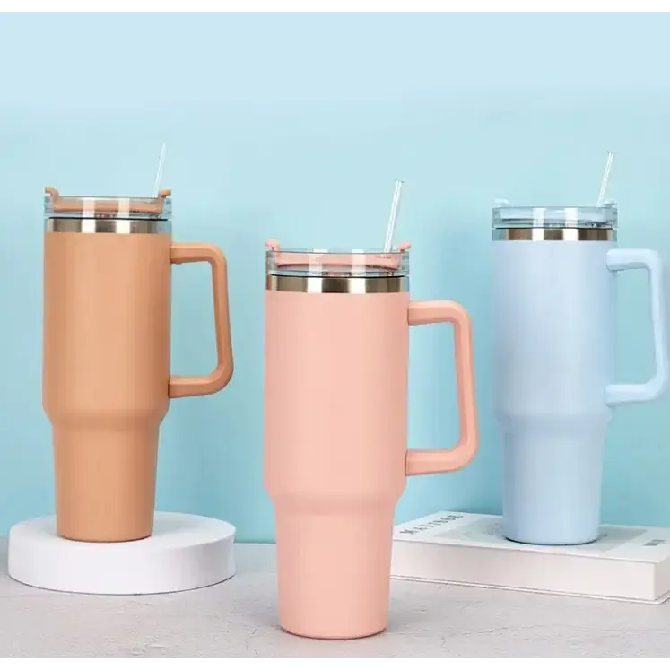 Three matte-finished insulated tumblers in soft peach, blush pink, and light blue hues, each featuring a sleek handle, clear lid with straw, and metallic rim.