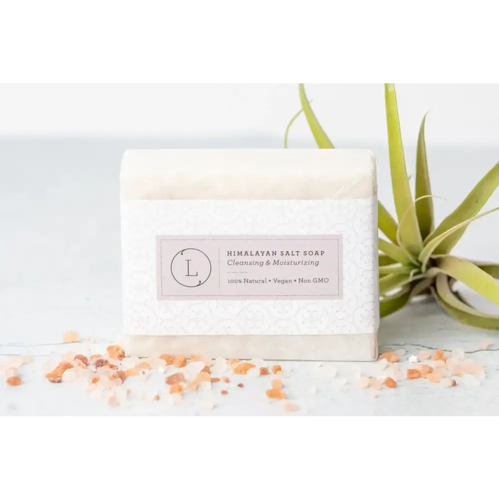 A rectangular bar of himalayan salt soap wrapped in a white patterned paper label with pink text, featuring the brand logo and descriptors like ’cleansing & moisturizing’ and ’100% natural • vegan • non gmo.’