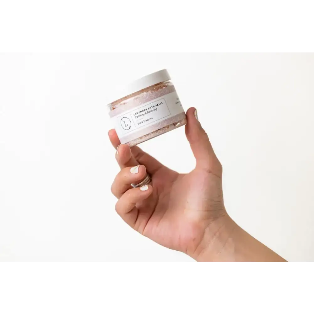 A clear glass jar with a white lid and label, containing lavender bath salts in pinkish-purple granules.