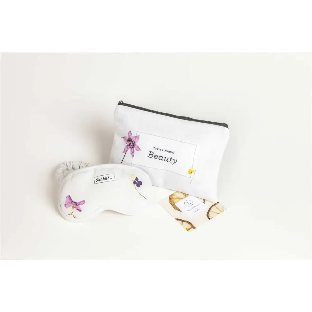 White eye mask with delicate purple floral embroidery and a small ’shhh...’ label, paired with a matching white pouch featuring the phrase “you’re a natural beauty” in black text.