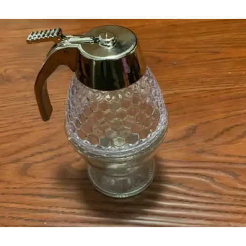 Clear glass honey dispenser with a metallic silver lid and handle.