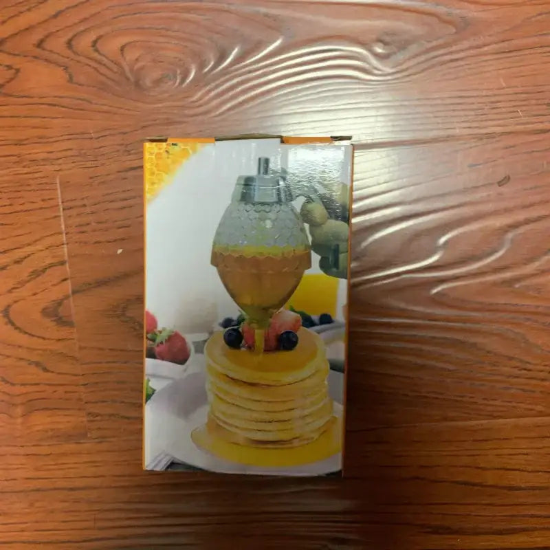 A clear glass honey dispenser with a silver metal top, designed to pour golden honey over pancakes.