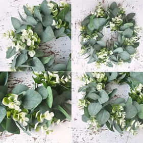 A circular wreath made of artificial eucalyptus leaves and small white flowers, featuring a mix of soft green and pale cream tones.