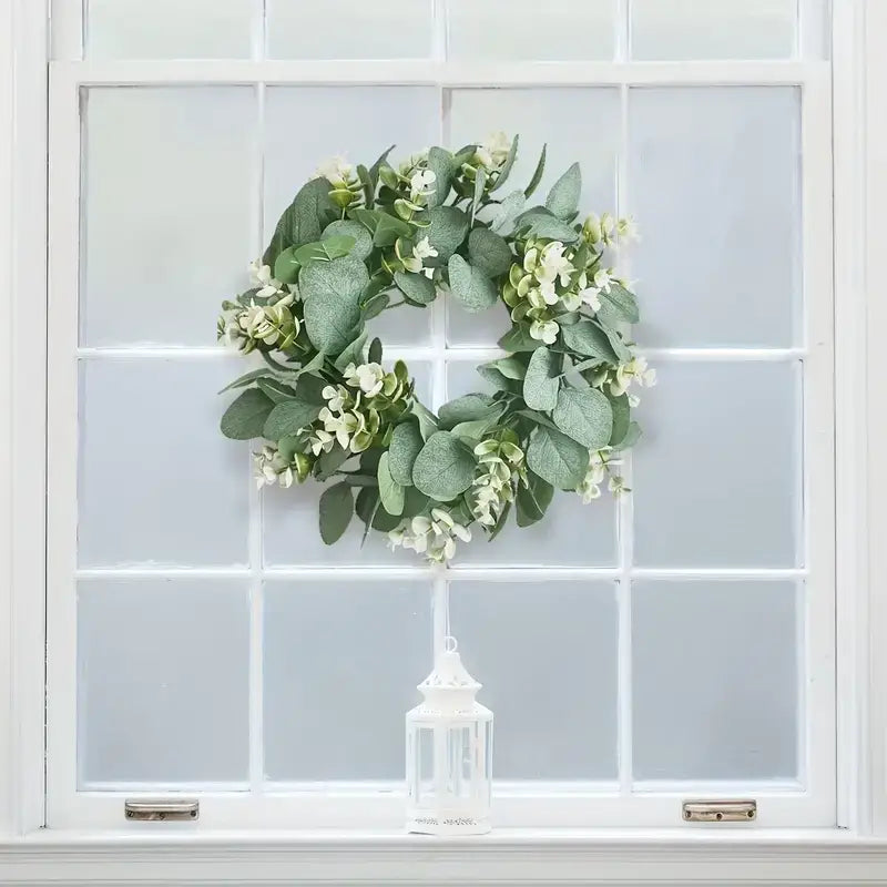 A lush green eucalyptus wreath adorned with delicate white flowers hangs gracefully on a window.