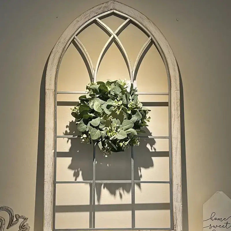 A rustic white-painted arched window frame with a decorative green eucalyptus wreath hanging centrally.