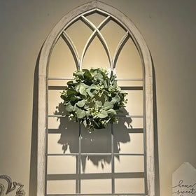 A rustic white-painted arched window frame with a decorative green eucalyptus wreath hanging centrally.