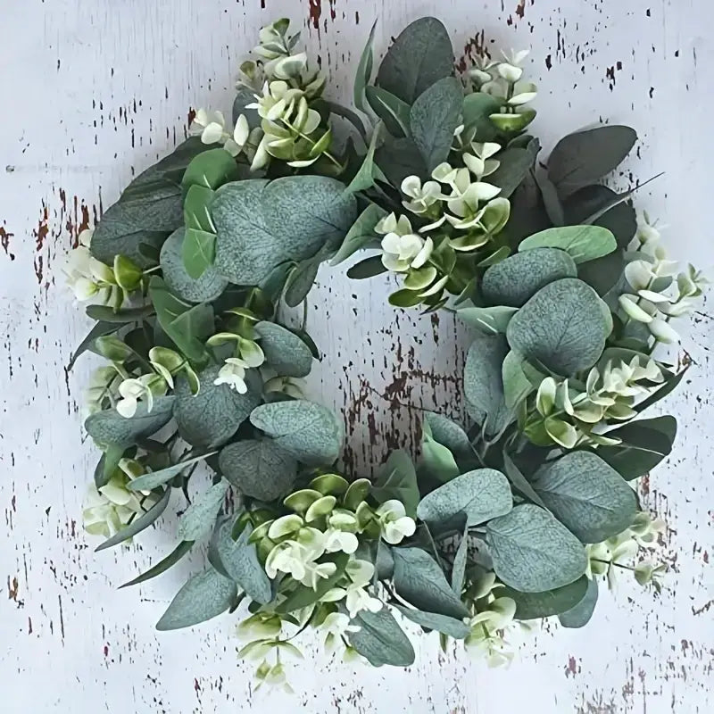A circular wreath made of artificial eucalyptus leaves and small white flowers, featuring a mix of muted green tones with subtle texture.
