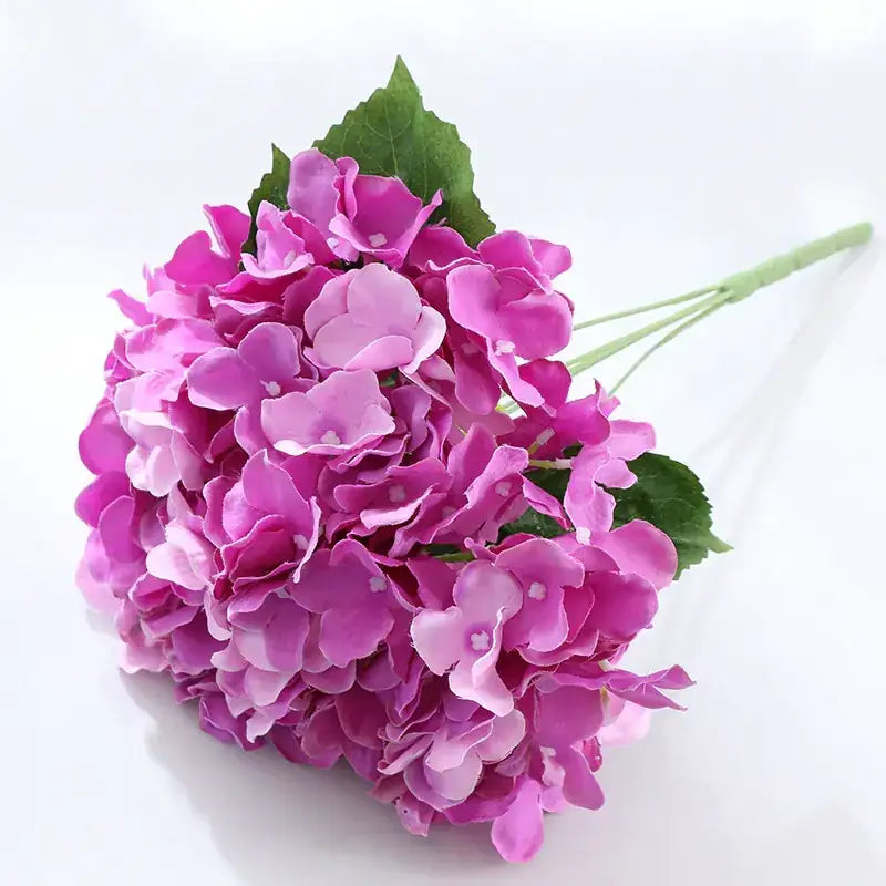 A vibrant bouquet of artificial hydrangeas with layered petals in shades of pink and purple, accented by green leaves.