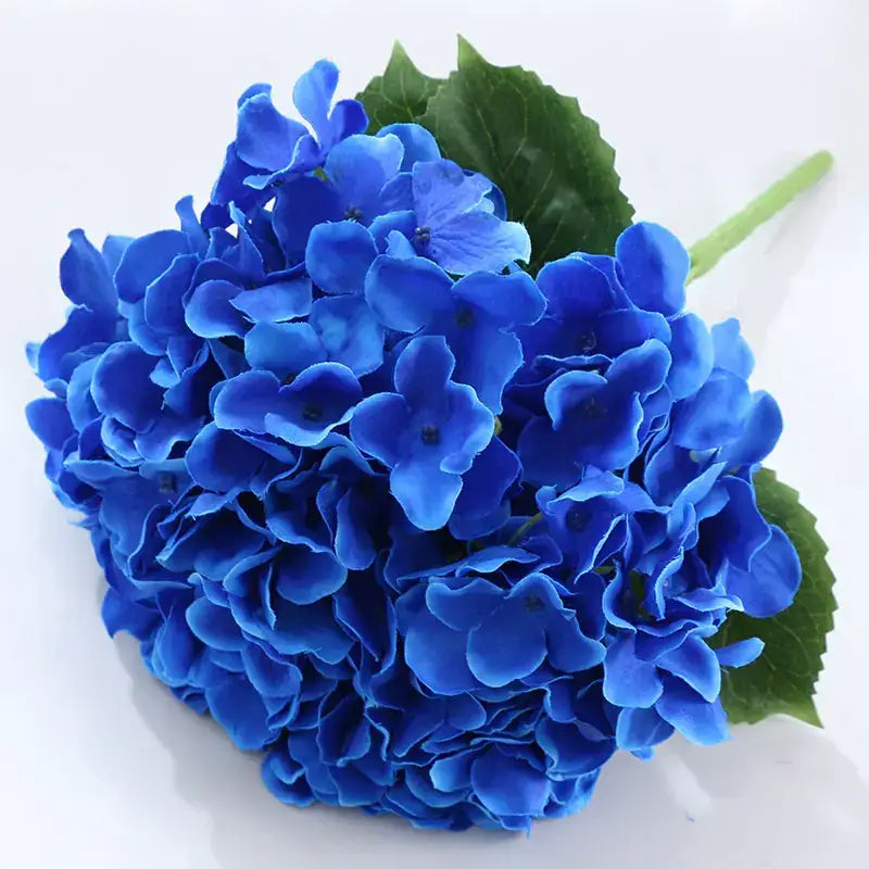 Vibrant royal blue artificial hydrangea bouquet with lush, layered petals and glossy green leaves.