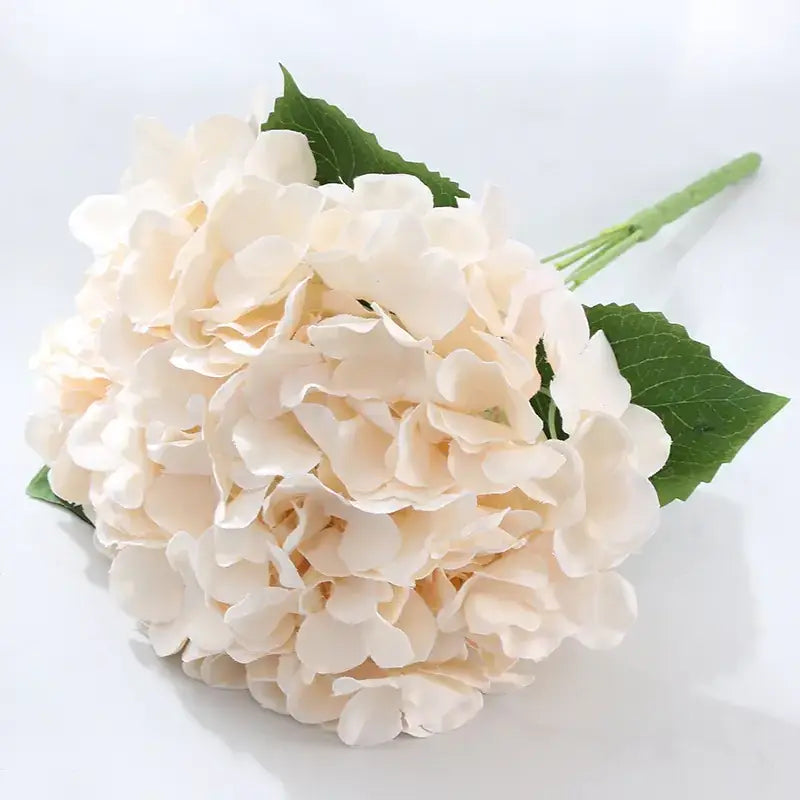 A delicate cream-colored artificial hydrangea bouquet with soft, layered petals and vibrant green leaves.