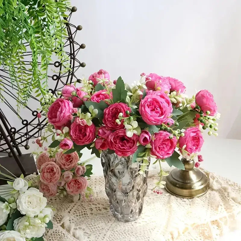 A clear, faceted glass vase holding a lush arrangement of vibrant pink and soft peach artificial roses with green leaves.