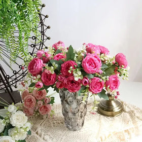 A clear, faceted glass vase holding a lush arrangement of vibrant pink and soft peach artificial roses with green leaves.