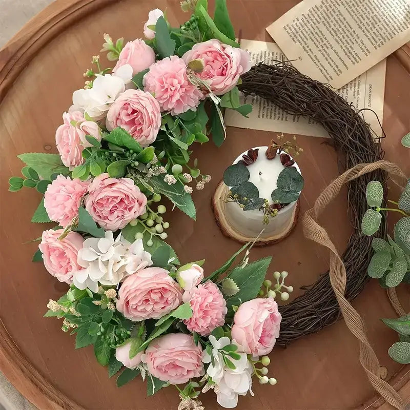 A delicate pink and white floral wreath adorned with lush greenery, crafted from artificial materials to mimic fresh blossoms.
