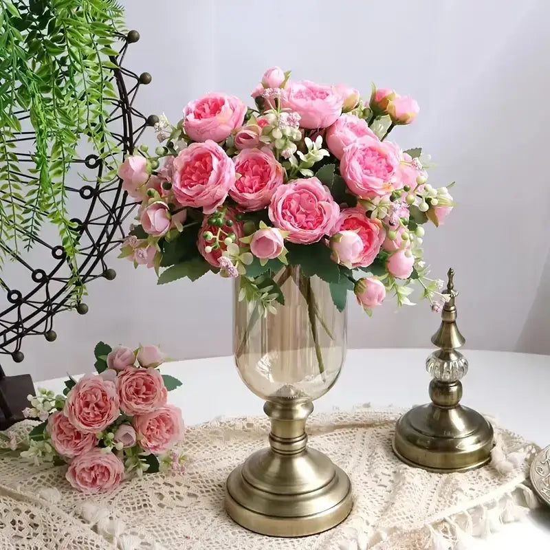 A tall, clear glass vase with a gold-toned metallic base and stem holds an arrangement of pink artificial roses.