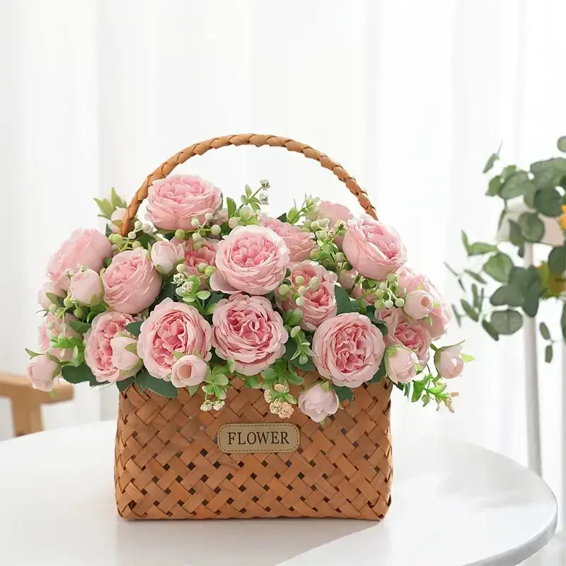 A woven wicker basket with a braided handle, filled to the brim with soft pink artificial roses and greenery.