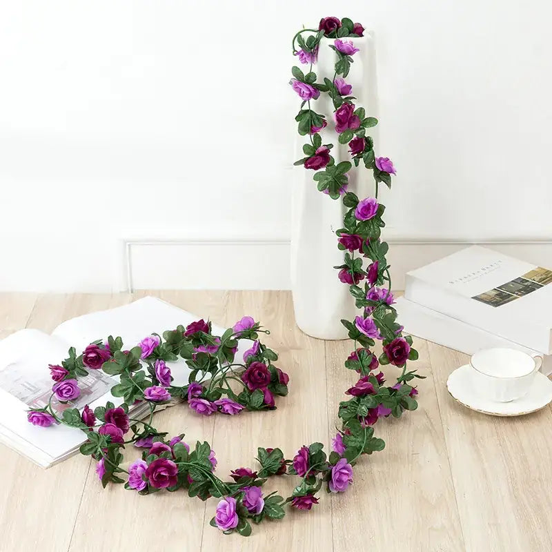 A decorative artificial rose garland with vibrant purple and deep magenta blooms intertwined with lush green leaves, crafted from synthetic material for a lifelike appearance.