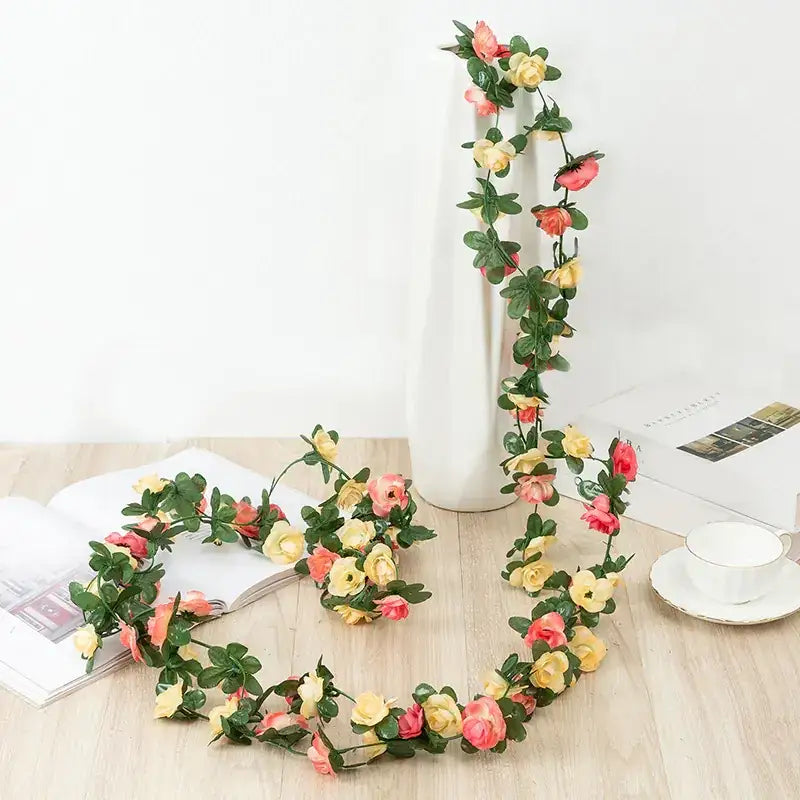 A delicate floral garland featuring artificial roses in soft pink and yellow hues with lush green leaves, crafted from synthetic materials for a realistic yet durable appearance.
