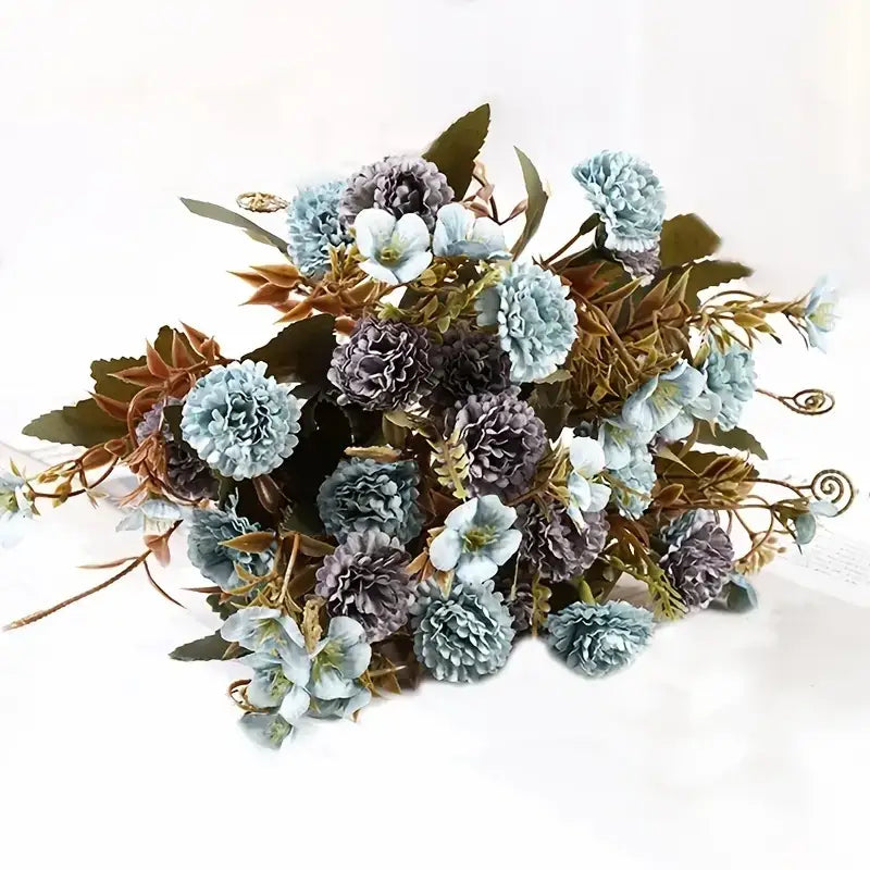 A delicate bouquet of artificial flowers in soft blue and muted purple hues, featuring textured petals and mixed greenery with subtle gold accents.