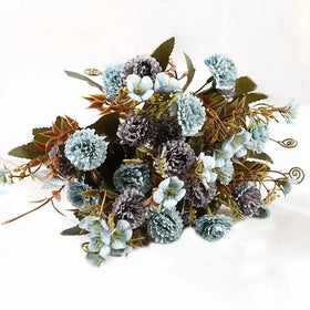 A delicate bouquet of artificial flowers in soft blue and muted purple hues, featuring textured petals and mixed greenery with subtle gold accents.