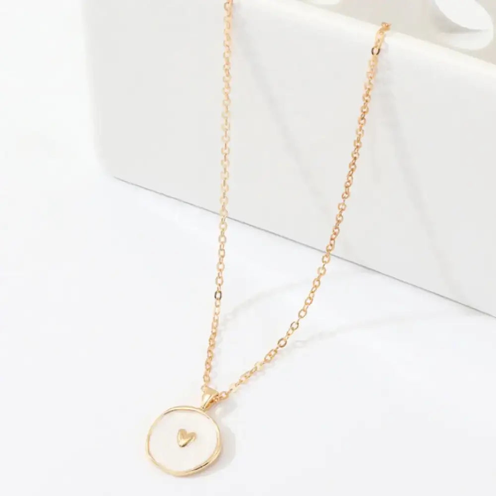 A delicate gold-toned necklace featuring a round, white enamel pendant with a small golden heart at its center.