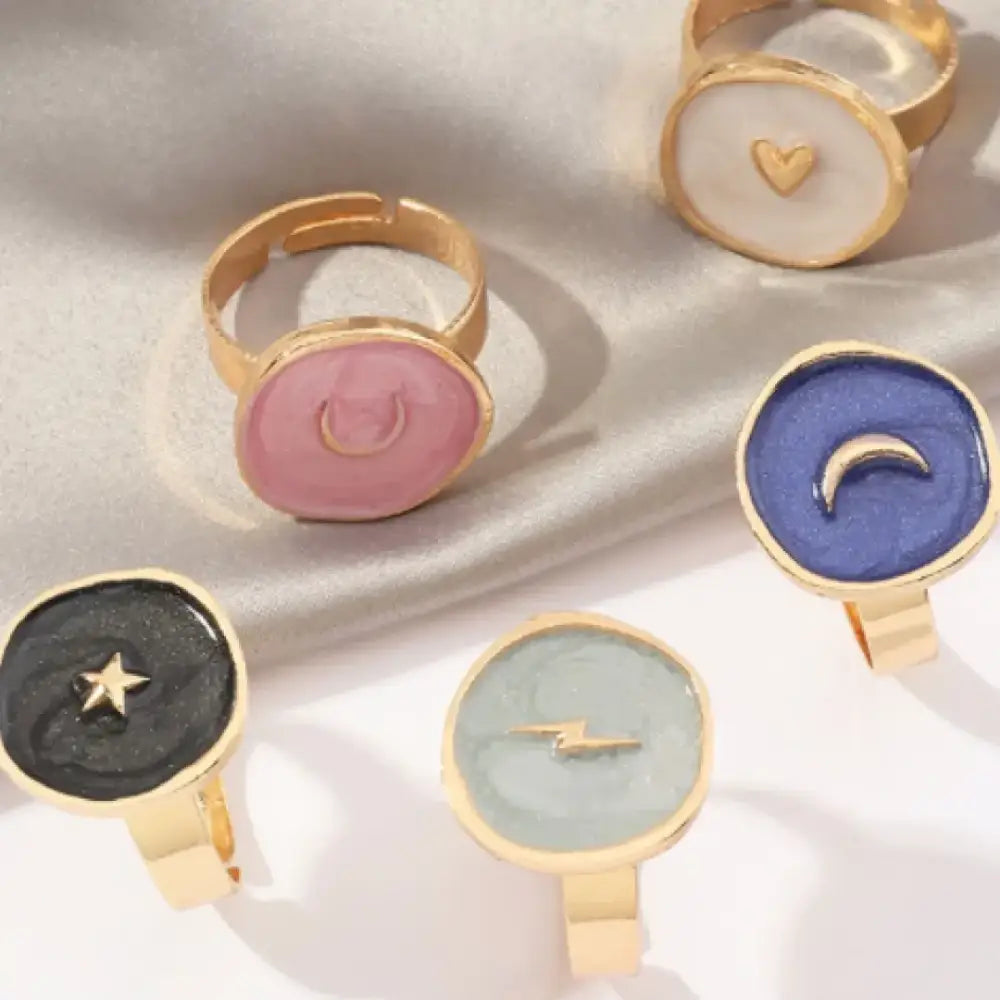 Gold-toned adjustable rings with flat, circular enamel faces featuring celestial and symbolic motifs in pastel pink, deep blue, black, and light green.