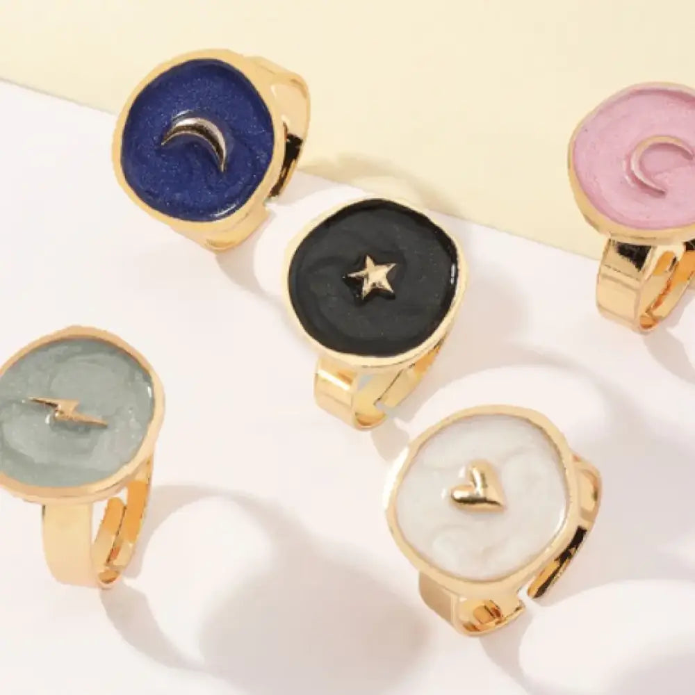 Gold-toned rings with flat, circular faces featuring colorful enamel designs—blue with a crescent moon, black with a star, pink with a swirl, gray-green with lightning, and white with a heart.