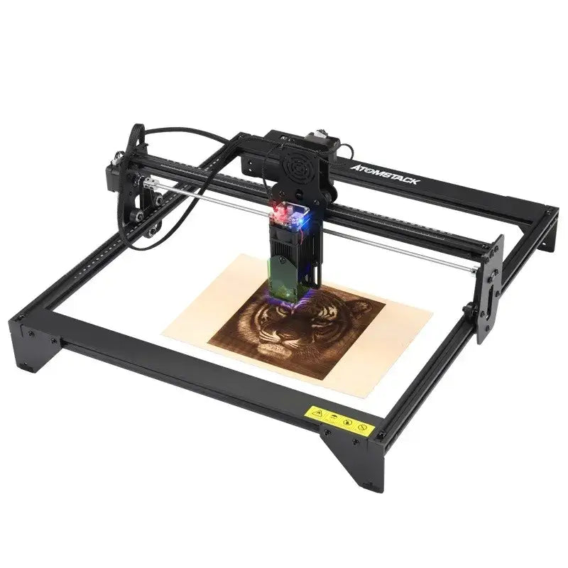 Black atomstack laser engraving machine with a glowing red and blue light head actively etching a tiger design onto beige material.