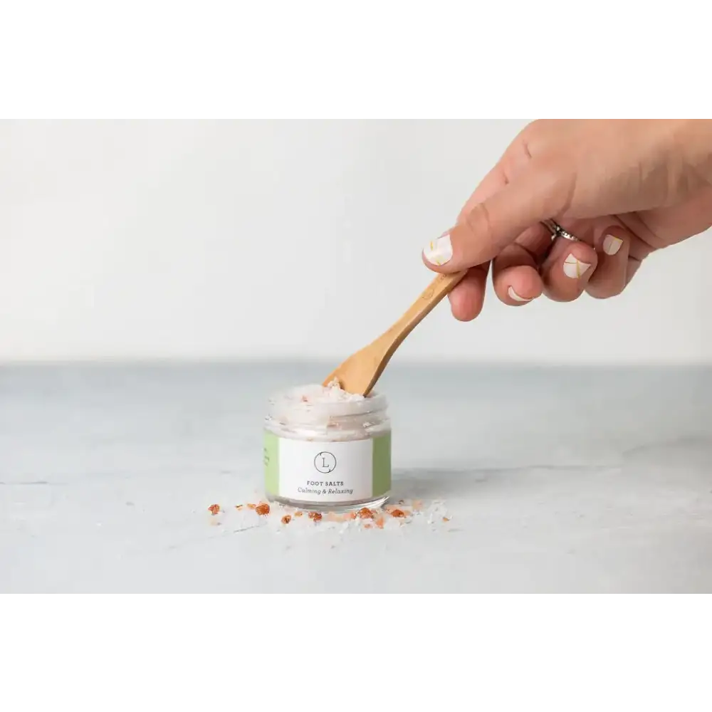 A clear glass jar with a green and white label containing pinkish-white foot salts, being stirred by a wooden spoon.