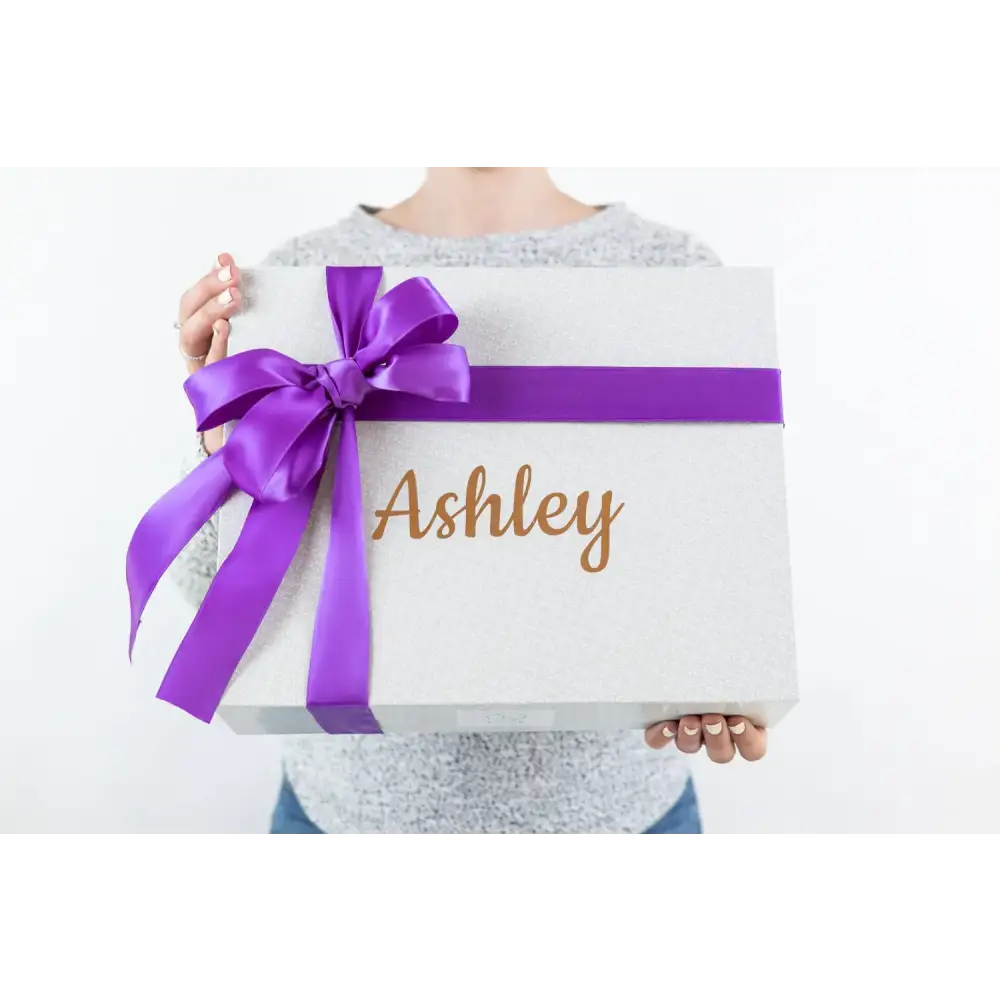 A white glittery gift box adorned with a vibrant purple satin ribbon bow and the name ’ashley’ in gold cursive lettering.